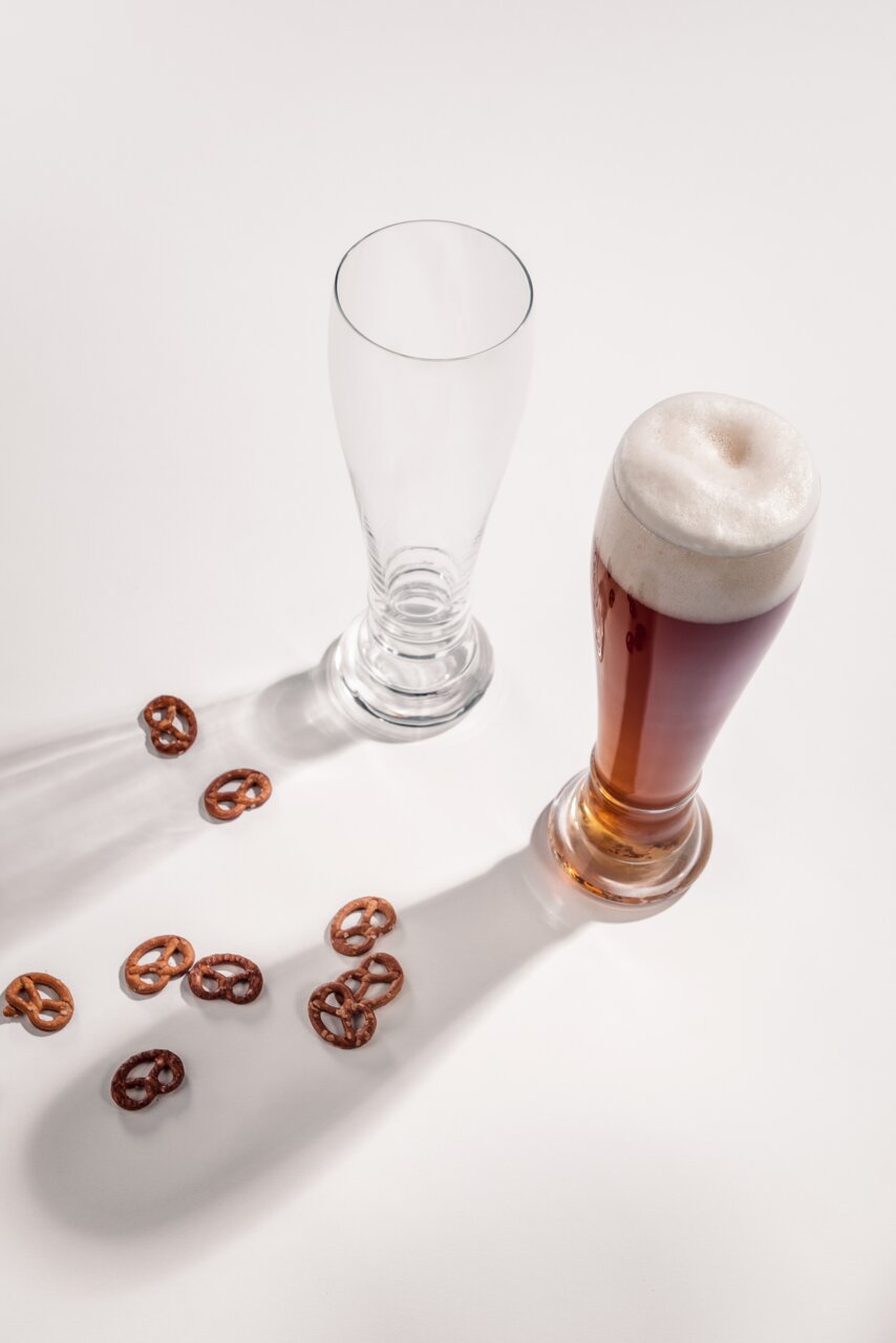 Twee glazen witbier, één leeg en één gevuld met donker bier, van bovenaf gefotografeerd, omringd door kleine pretzels op een witte achtergrond.