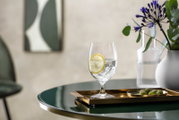 Ein eleganter Wasserkelch mit Eiswürfeln und einer Zitronenscheibe, auf einem goldenen Tablett auf einem Tisch, umgeben von einer stilvollen Dekoration. Perspektive: seitlich.