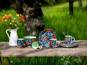 Buntes Geschirrset mit floralen Mustern (Tassen, Schalen und Teller) auf rustikalem Holztisch im Garten neben weißer Vase mit Blumen, Perspektive: frontale Tischhöhe/leichte Augenhöhe.