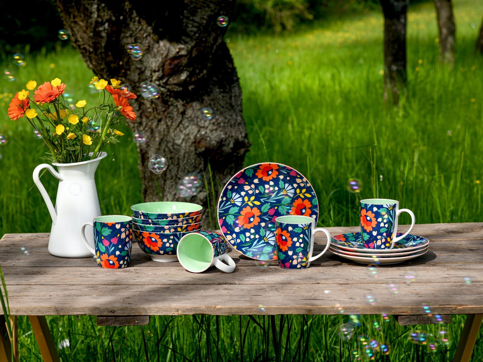 Buntes Geschirrset mit floralen Mustern (Tassen, Schalen und Teller) auf rustikalem Holztisch im Garten neben weißer Vase mit Blumen, Perspektive: frontale Tischhöhe/leichte Augenhöhe.