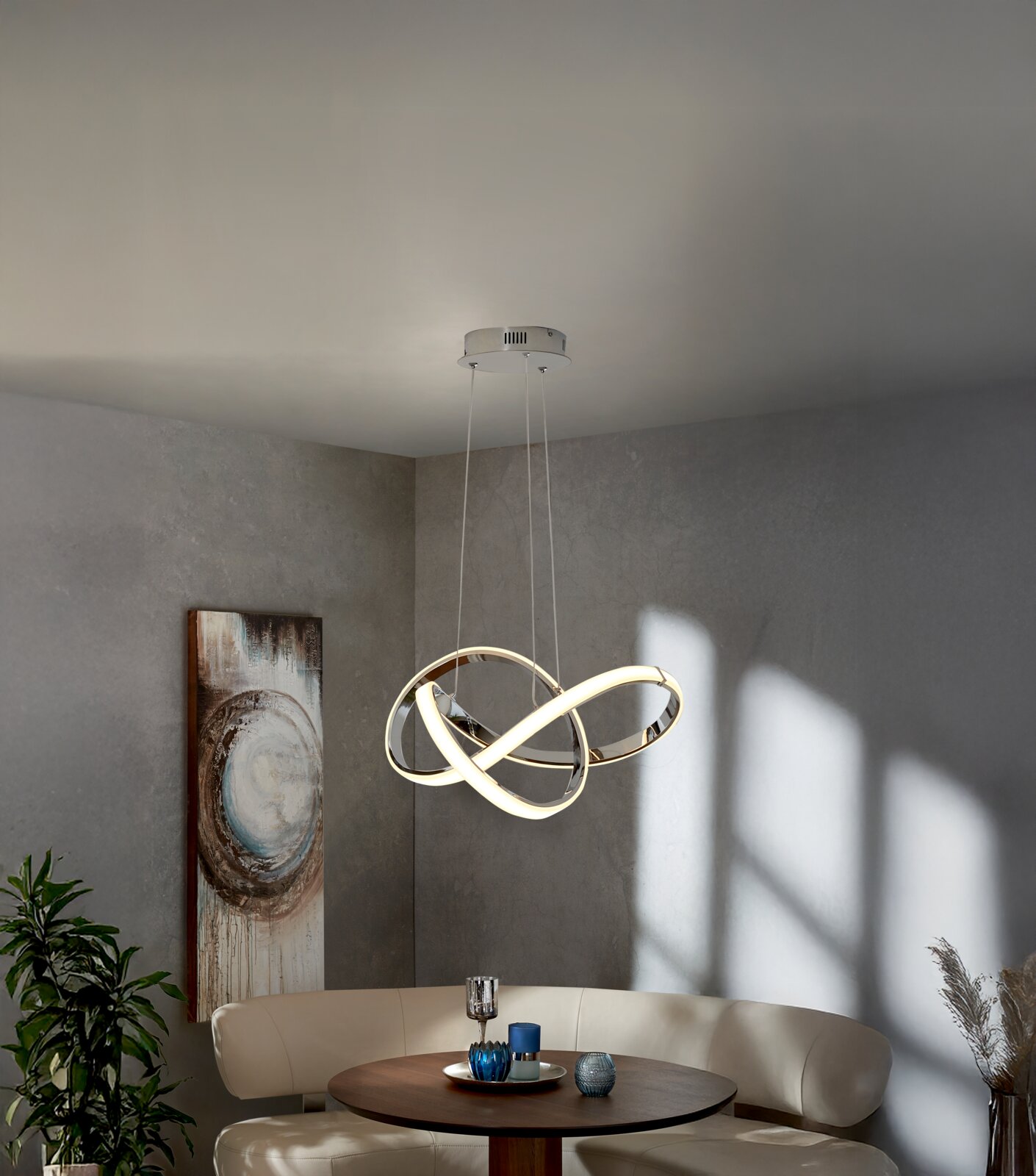Moderne LED-Pendelleuchte mit verschlungenen, ringförmigen Lichtbändern in Chromoptik über einem runden Esstisch in grauem Wohnraum, Ansicht leicht von unten (Untersicht)