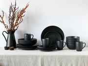 Schwarzes Kombiservice Soft Touch 20-tlg, bestehend aus Tellern, Schalen und Tassen, auf einem Tisch mit Leinentuch, seitliche Perspektive.