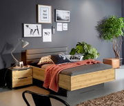 Modern houten bed met een grijs gestoffeerd hoofdeinde in een stijlvolle slaapkamer, met nachtkastje en lamp, grijs sprei en rode deken, wanddecoraties en kamerplanten; perspectief: schuin van linksvoor.