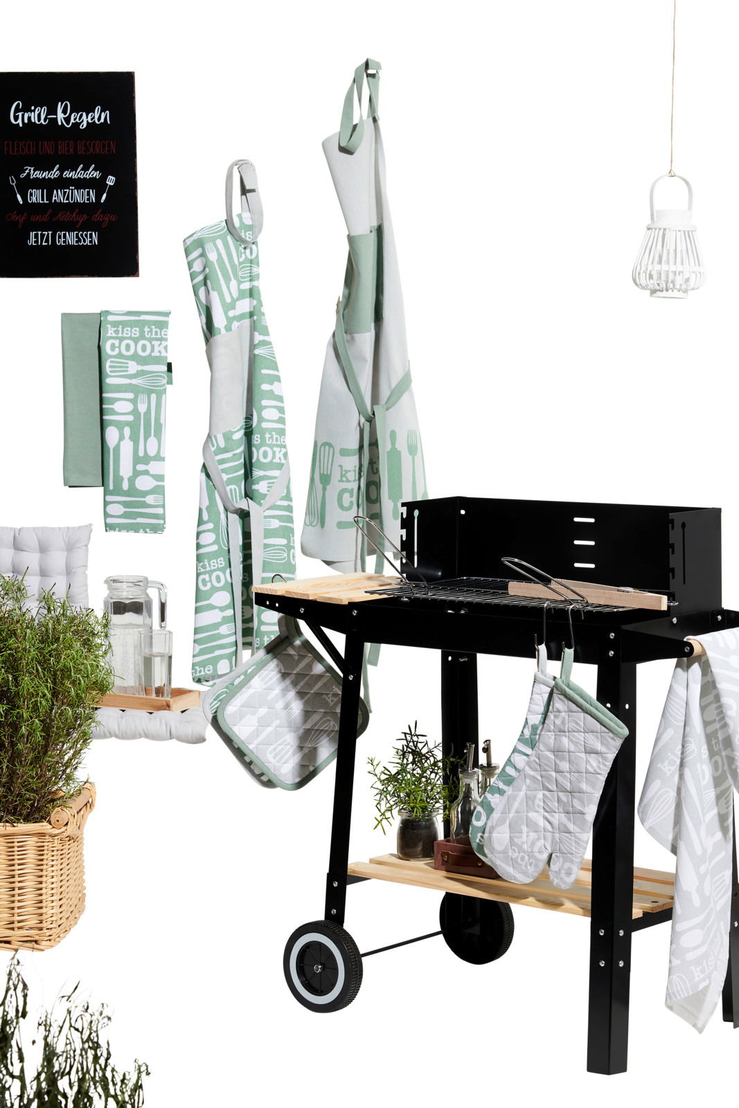Küchen- und Grillzubehör-Set mit Geschirrtüchern, Schürzen und Topflappen in grün-weißem Design mit der Aufschrift 'Kiss the Cook', präsentiert neben einem Grill aus seitlicher Perspektive.