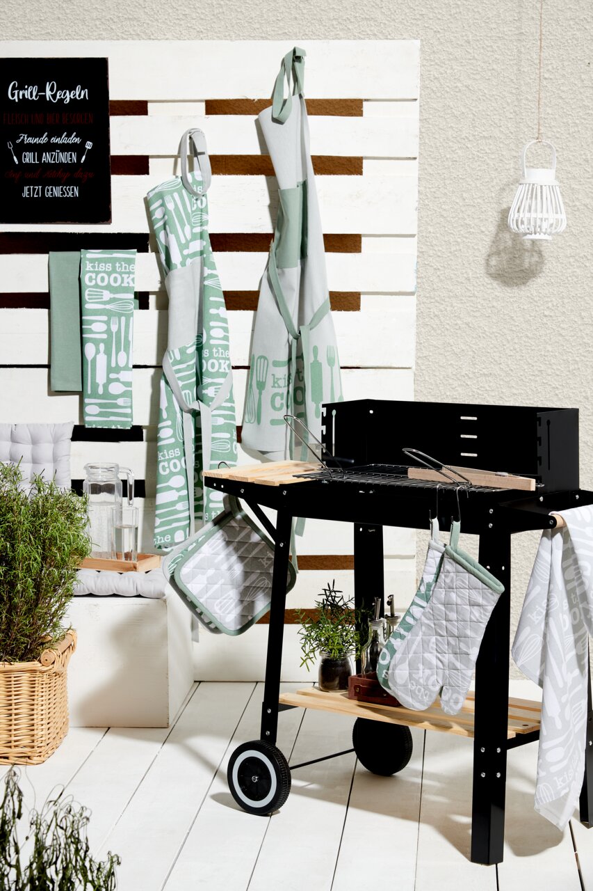 Küchen- und Grillzubehör-Set mit Geschirrtüchern, Schürzen und Topflappen in grün-weißem Design mit der Aufschrift 'Kiss the Cook', präsentiert neben einem Grill aus seitlicher Perspektive.