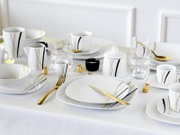 Schickes weißes Porzellan-Tafelservice mit schwarzen und goldenen Linien (Teller, Schalen, Tassen, Kanne und Zuckerdose) auf gedecktem Tisch mit goldfarbenem Besteck, fotografiert aus leicht erhöhter, schräger Frontperspektive.