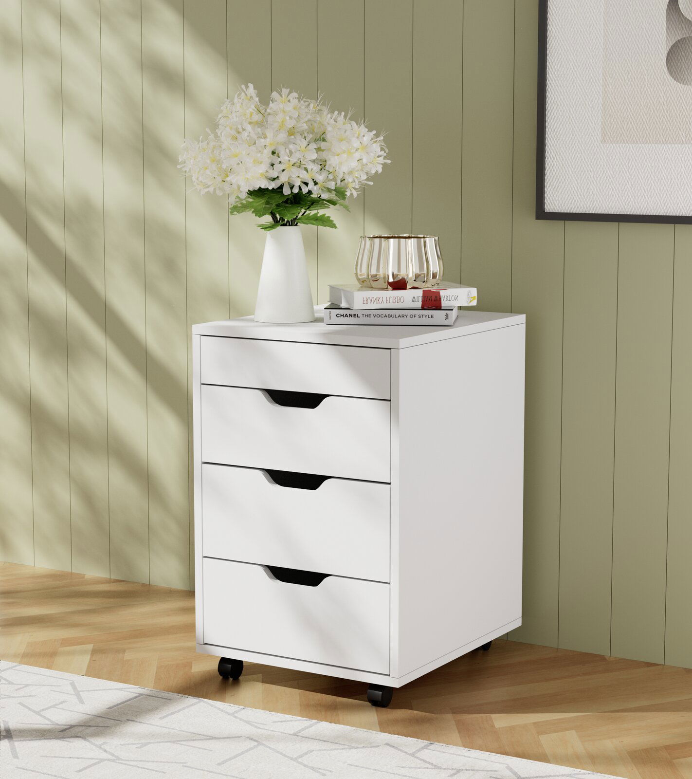 Carryhome Rollcontainer JULIUS Weißer Rollcontainer mit drei Schubladen, dekoriert mit einer Vase voller Blumen und Büchern, auf einem Holzboden vor einer grünen Wand. Perspektive von vorne links.