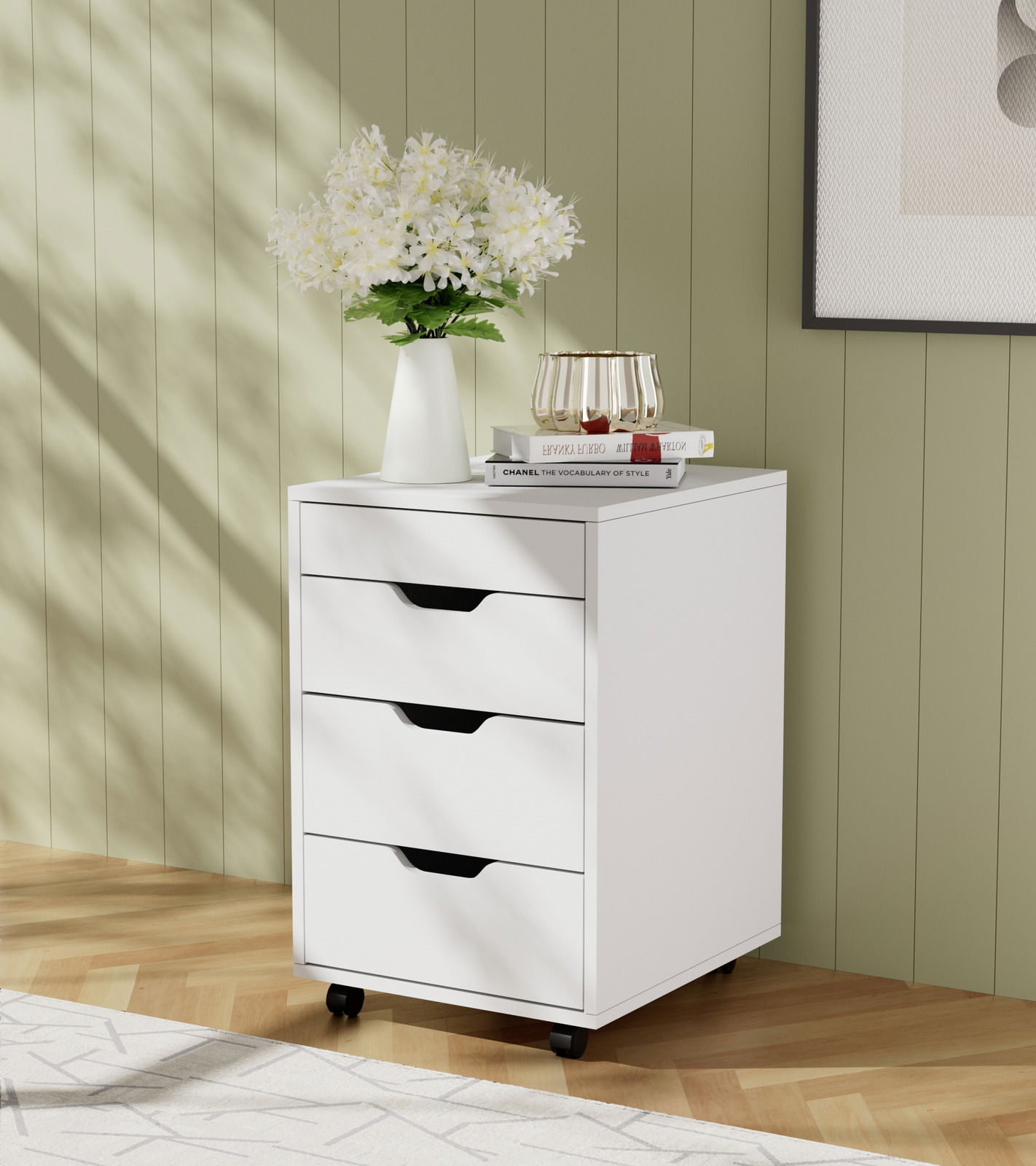 Weißer Rollcontainer mit drei Schubladen, dekoriert mit einer Vase voller Blumen und Büchern, auf einem Holzboden vor einer grünen Wand. Perspektive von vorne links.