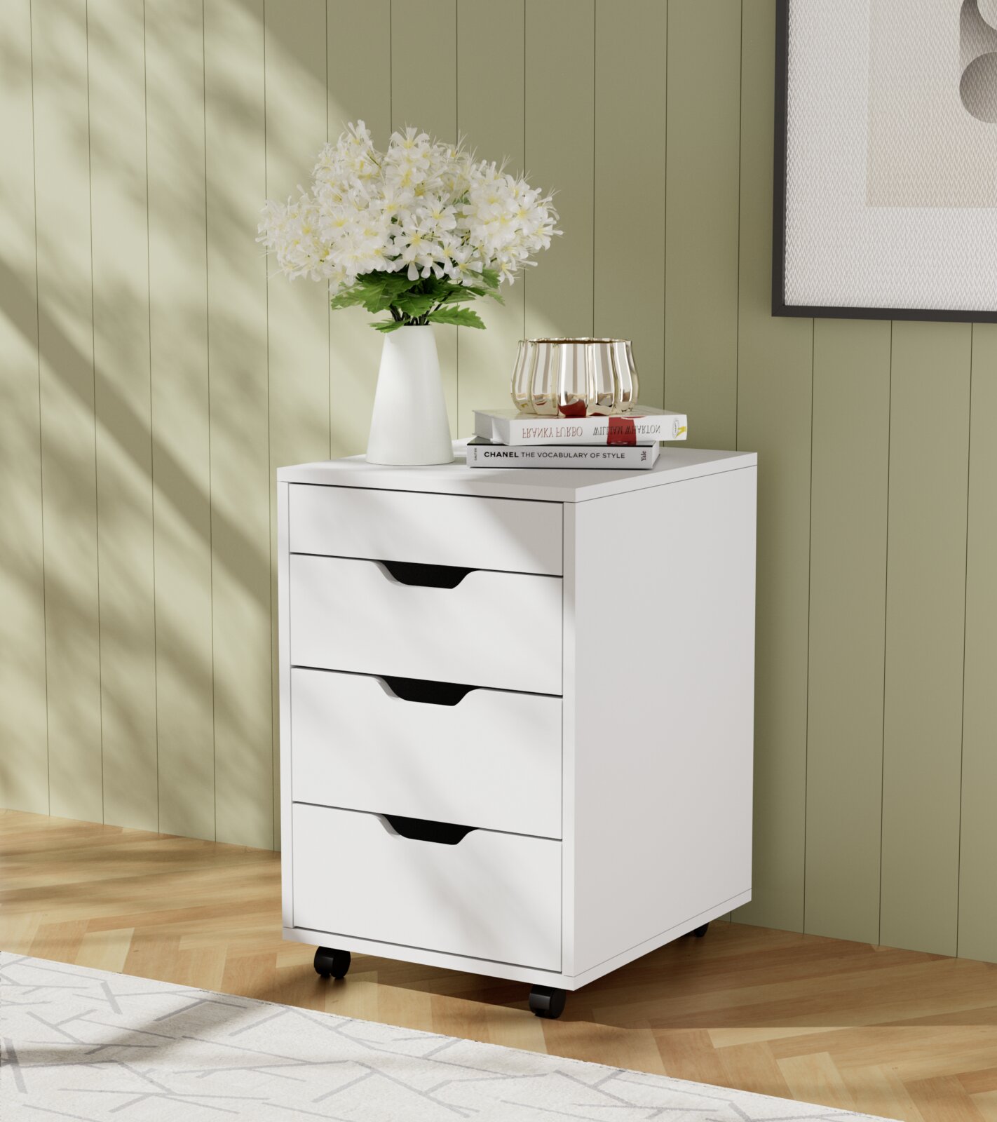 Weißer Rollcontainer mit drei Schubladen, dekoriert mit einer Vase voller Blumen und Büchern, auf einem Holzboden vor einer grünen Wand. Perspektive von vorne links.