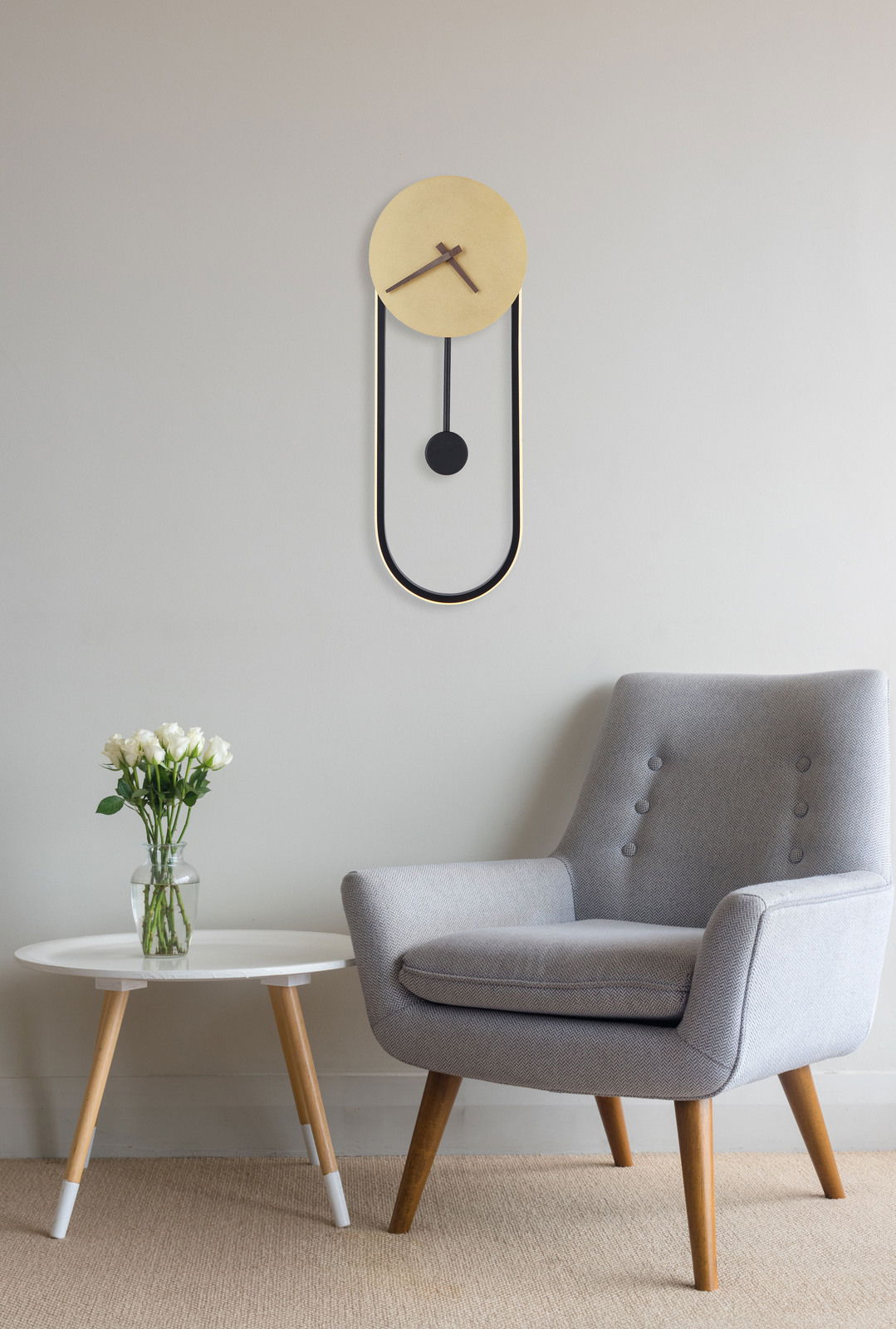 Moderne Wanduhr SUSSY in minimalistischem Design, goldener Ziffernblatt mit schwarzem Pendel, an einer hellen Wand über einem grauen Sessel und einem weißen Beistelltisch mit Blumen, aus der Seitenperspektive.