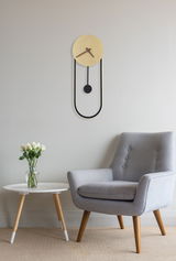 Moderne Wanduhr SUSSY in minimalistischem Design, goldener Ziffernblatt mit schwarzem Pendel, an einer hellen Wand über einem grauen Sessel und einem weißen Beistelltisch mit Blumen, aus der Seitenperspektive.