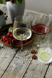 Zwei Weingläser, eines mit Rotwein und eines mit Weißwein, auf einem Holztisch mit Früchten und Gewürzen im Hintergrund, aus einer leicht erhöhten Perspektive fotografiert.