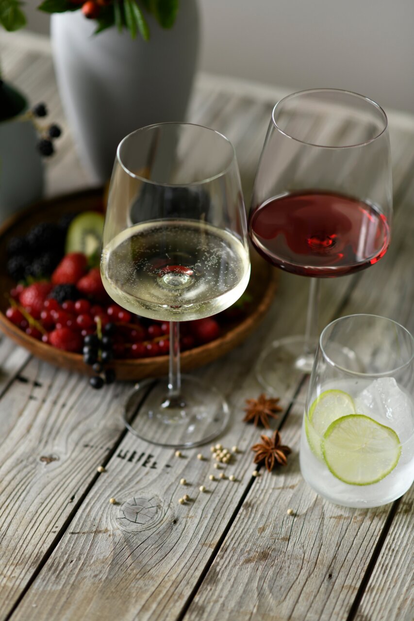 Zwei Weingläser, eines mit Rotwein und eines mit Weißwein, auf einem Holztisch mit Früchten und Gewürzen im Hintergrund, aus einer leicht erhöhten Perspektive fotografiert.