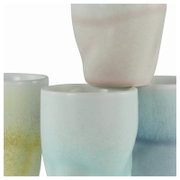 Stapel aus glasierten Keramikbechern in Pastellfarben (Gelb, Mint, Blau und Rosé) vor weißem Hintergrund, Nahaufnahme aus leicht seitlicher Perspektive.