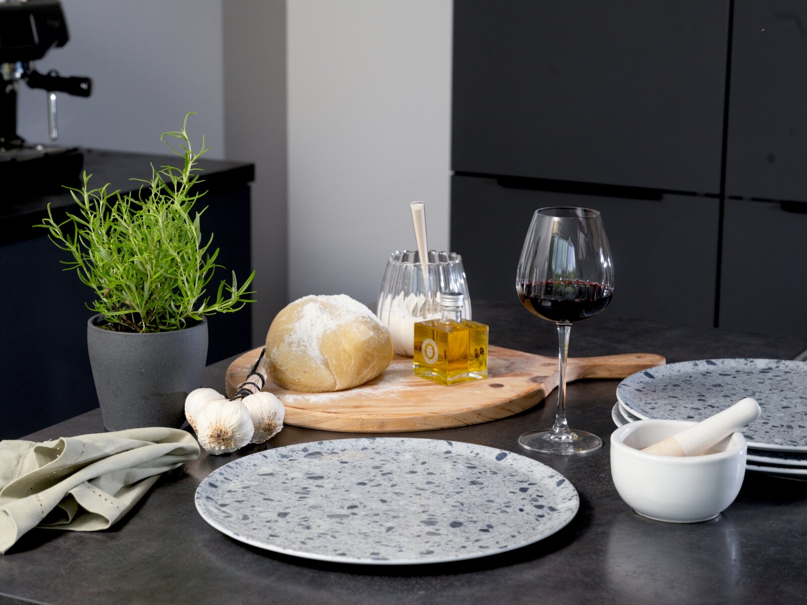 Modernes Tisch-Setting in einer dunklen Küche: graue, gesprenkelte Keramikteller auf dem Tisch, daneben weißer Mörser, Rotweinglas, Brot auf Holzbrett mit Olivenöl, Knoblauch und Kräutertopf – Perspektive: frontale Tischhöhe mit leicht seitlichem Blick vo