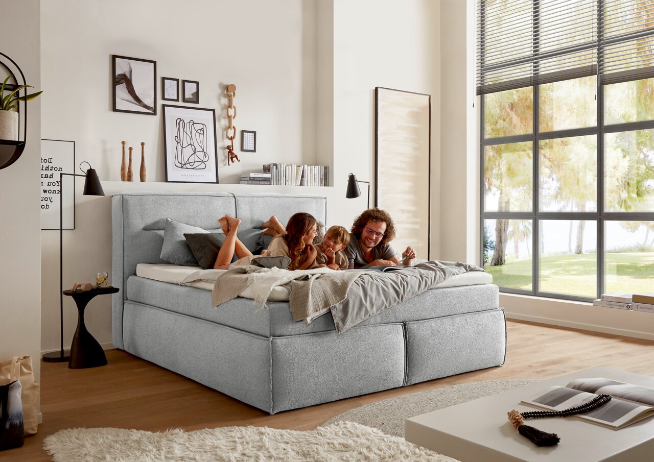Gemütliches Boxspringbett in einem modernen Schlafzimmer mit großen Fenstern, von der Seite betrachtet. Eine Familie liegt entspannt auf dem Bett.