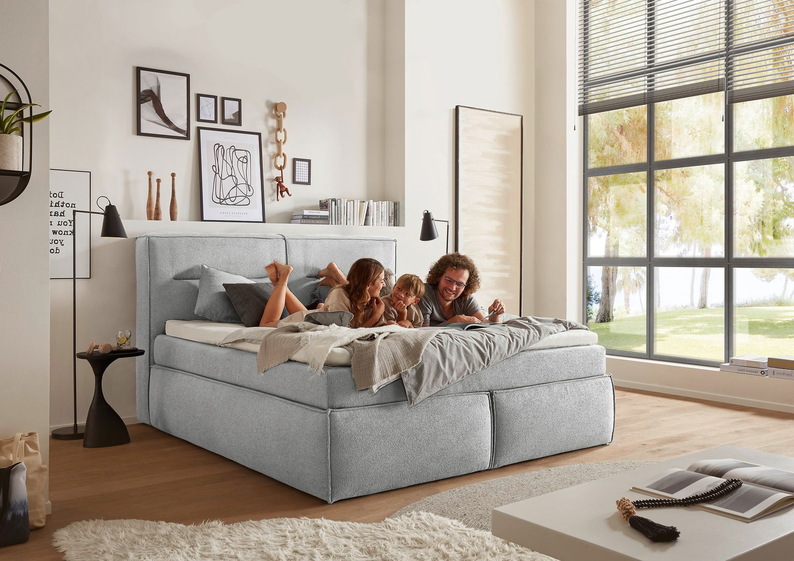Gemütliches Boxspringbett in einem modernen Schlafzimmer mit großen Fenstern, von der Seite betrachtet. Eine Familie liegt entspannt auf dem Bett.
