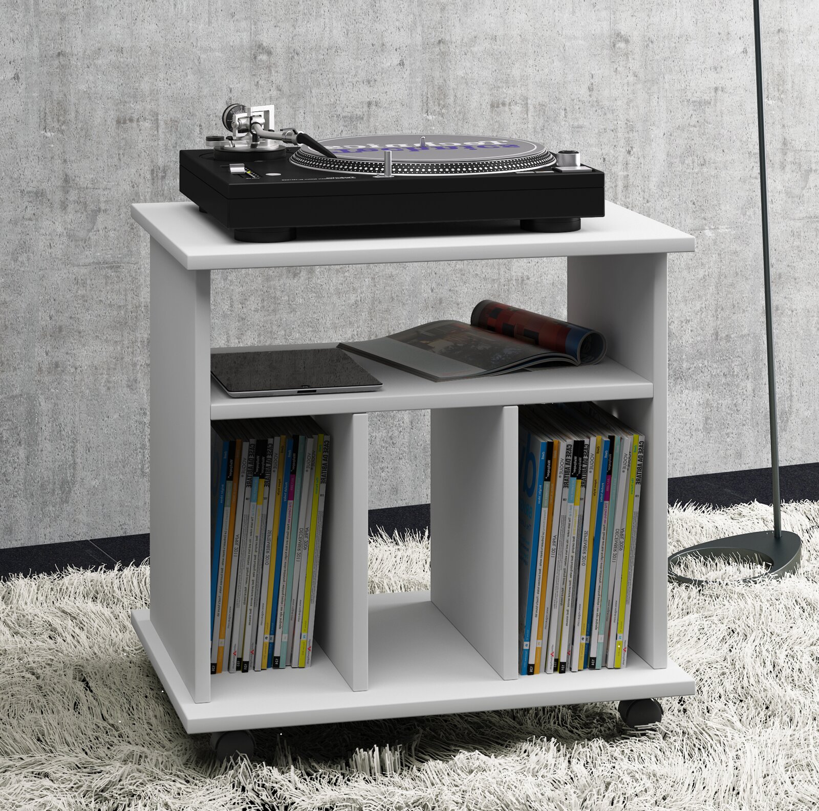 Weißes, modernes Regal-/Beistelltisch-Modul auf Rollen mit offenen Fächern und Zeitschriften, darauf ein schwarzer Plattenspieler; Ansicht schräg von vorne.