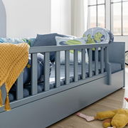 Nahaufnahme eines grauen Bodenbetts mit Rausfallschutz, dekoriert mit bunten Kissen und einer gelben Decke, seitliche Perspektive.