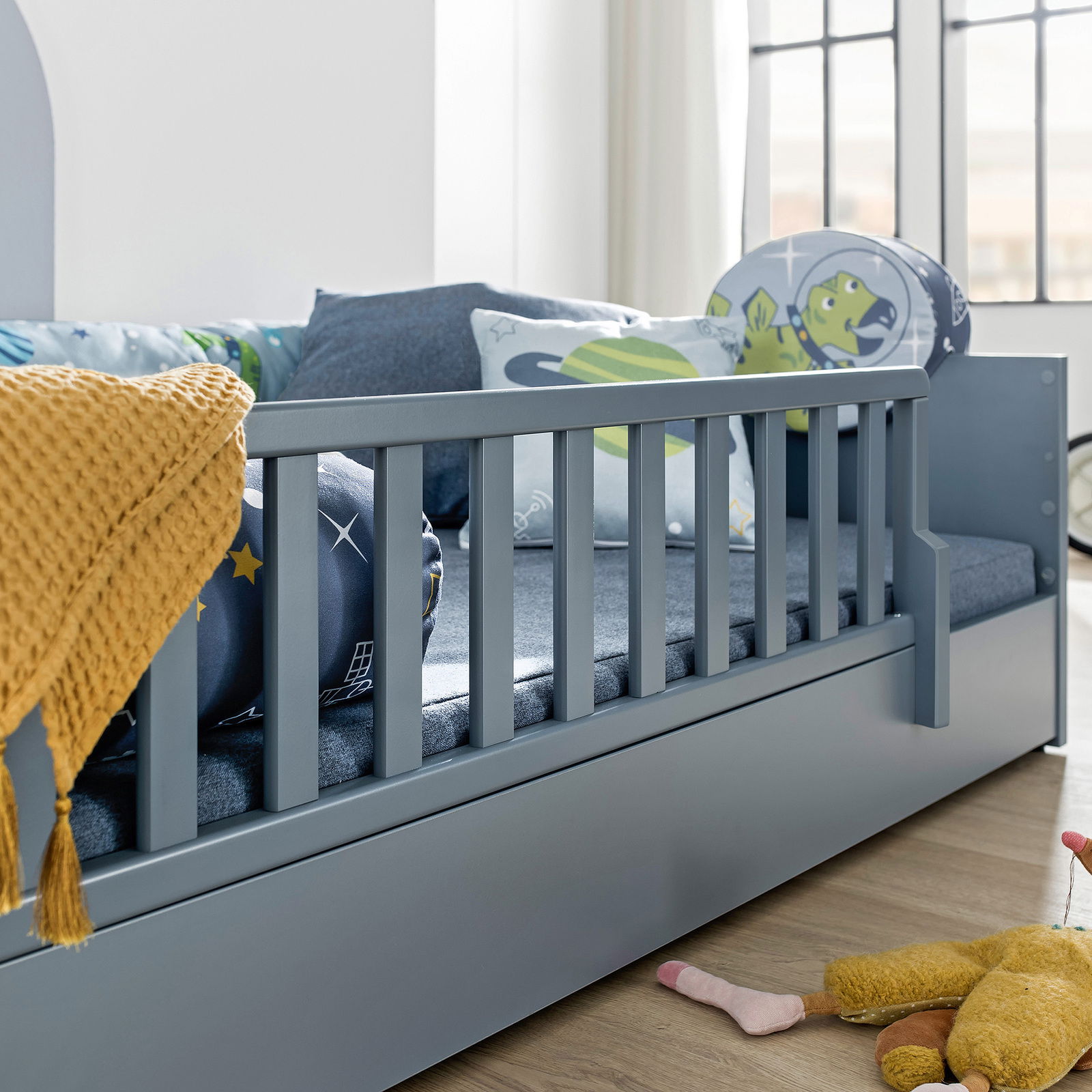 Nahaufnahme eines grauen Bodenbetts mit Rausfallschutz, dekoriert mit bunten Kissen und einer gelben Decke, seitliche Perspektive.