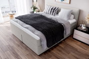 Modernes graues Boxspringbett mit gepolstertem Kopfteil, grauer Bettwäsche und schwarzer grob gestrickter Tagesdecke im hellen Schlafzimmer, aufgenommen aus schräger Vorderansicht von links oben.