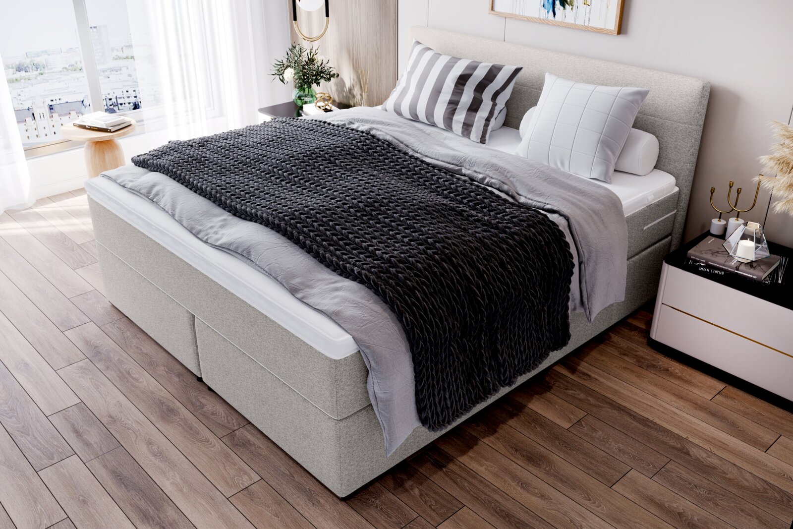 Modernes graues Boxspringbett mit gepolstertem Kopfteil, grauer Bettwäsche und schwarzer grob gestrickter Tagesdecke im hellen Schlafzimmer, aufgenommen aus schräger Vorderansicht von links oben.