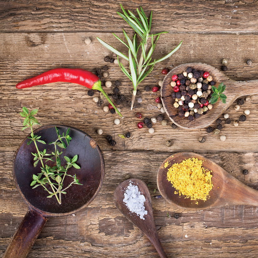 Vogelperspektive auf eine rustikale Holzoberfläche mit verschiedenen Kräutern und Gewürzen, darunter ein roter Chili, Rosmarin, Pfefferkörner, Thymian, grobes Salz und gelbes Gewürzpulver in Holzlöffeln.