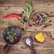 Vogelperspektive auf eine rustikale Holzoberfläche mit verschiedenen Kräutern und Gewürzen, darunter ein roter Chili, Rosmarin, Pfefferkörner, Thymian, grobes Salz und gelbes Gewürzpulver in Holzlöffeln.