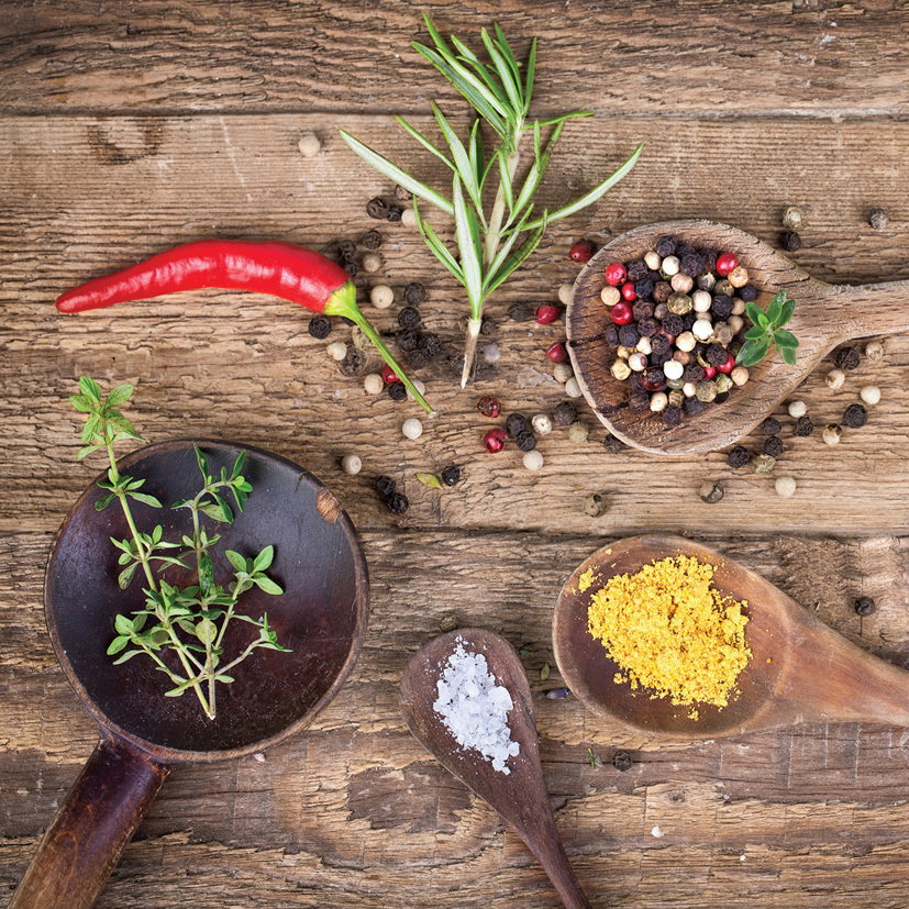 Vogelperspektive auf eine rustikale Holzoberfläche mit verschiedenen Kräutern und Gewürzen, darunter ein roter Chili, Rosmarin, Pfefferkörner, Thymian, grobes Salz und gelbes Gewürzpulver in Holzlöffeln.