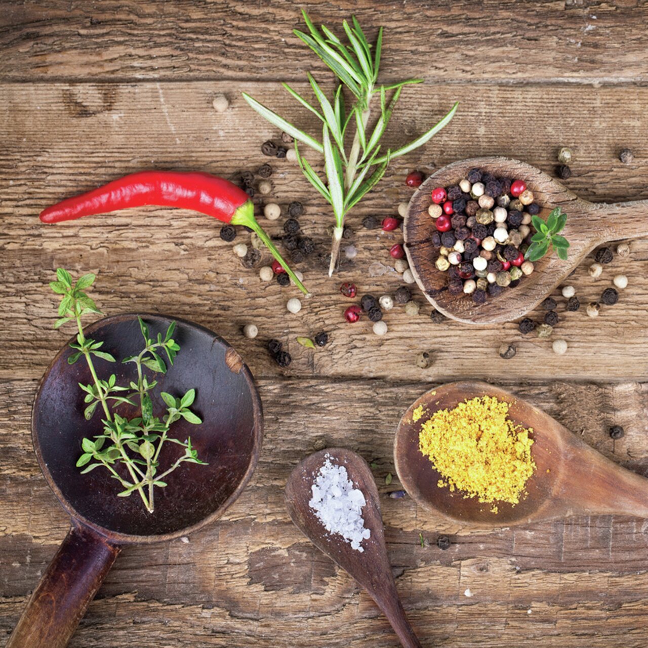 Vogelperspektive auf eine rustikale Holzoberfläche mit verschiedenen Kräutern und Gewürzen, darunter ein roter Chili, Rosmarin, Pfefferkörner, Thymian, grobes Salz und gelbes Gewürzpulver in Holzlöffeln.