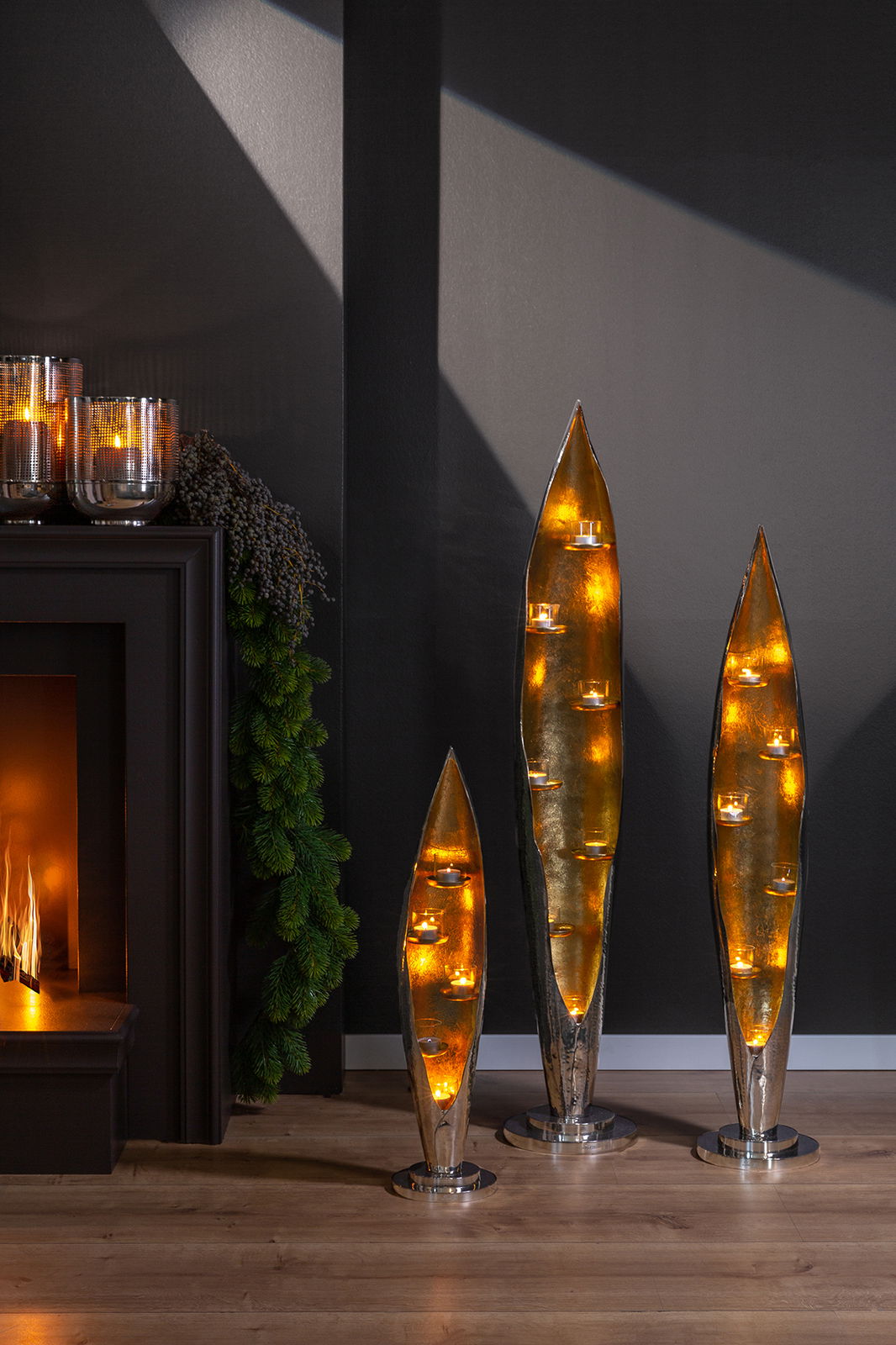 Drei moderne, goldene Kerzenhalter in Blattform mit Teelichtern, stehend auf einem Holzfußboden vor einer dunklen Wand. Perspektive: Frontalansicht.