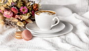 Weiße Kaffeetasse mit Untertasse auf einem weißen Tuch, begleitet von zwei Macarons und einem Blumenstrauß, seitliche Perspektive