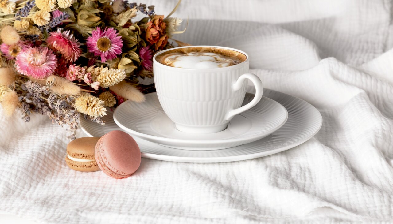 Weiße Kaffeetasse mit Untertasse auf einem weißen Tuch, begleitet von zwei Macarons und einem Blumenstrauß, seitliche Perspektive
