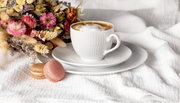 Witte koffiekop met schoteltje op een wit kleed, vergezeld van twee macarons en een boeket bloemen, zijaanzicht