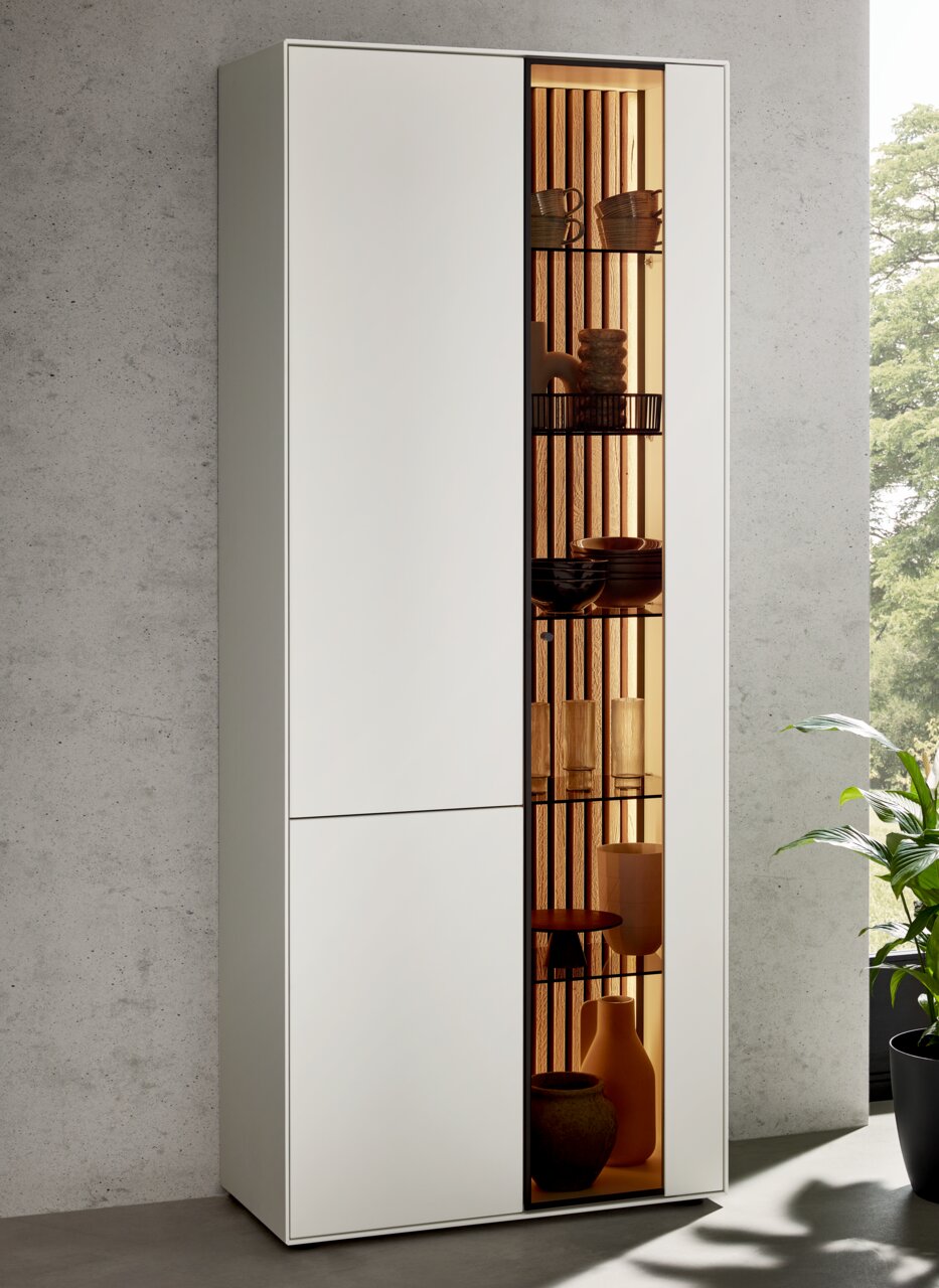 Moderne Standvitrine LUCIANO mit linksseitiger Tür und rechtsseitigem Glaseinsatz, frontal betrachtet, mit dekorativer Beleuchtung und stilvollen Accessoires.