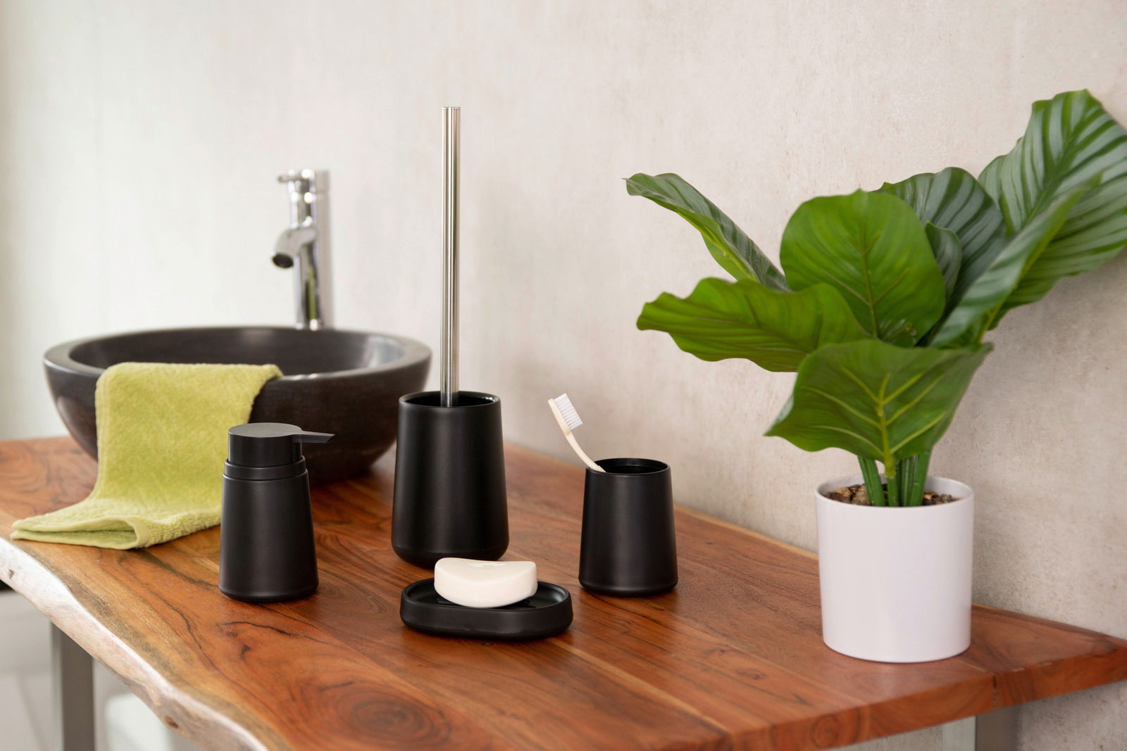 Badezimmer-Set Kira in Schwarz, bestehend aus Seifenspender, WC-Bürste, Seifenschale und Aufbewahrungsbecher, auf einem Holztisch neben einem Waschbecken und einer Pflanze, seitliche Perspektive.