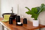 Badezimmer-Set Kira in Schwarz, bestehend aus Seifenspender, WC-Bürste, Seifenschale und Aufbewahrungsbecher, auf einem Holztisch neben einem Waschbecken und einer Pflanze, seitliche Perspektive.