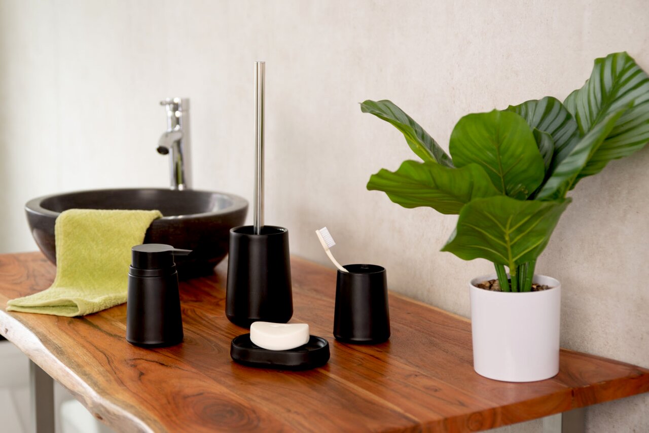 Badezimmer-Set Kira in Schwarz, bestehend aus Seifenspender, WC-Bürste, Seifenschale und Aufbewahrungsbecher, auf einem Holztisch neben einem Waschbecken und einer Pflanze, seitliche Perspektive.