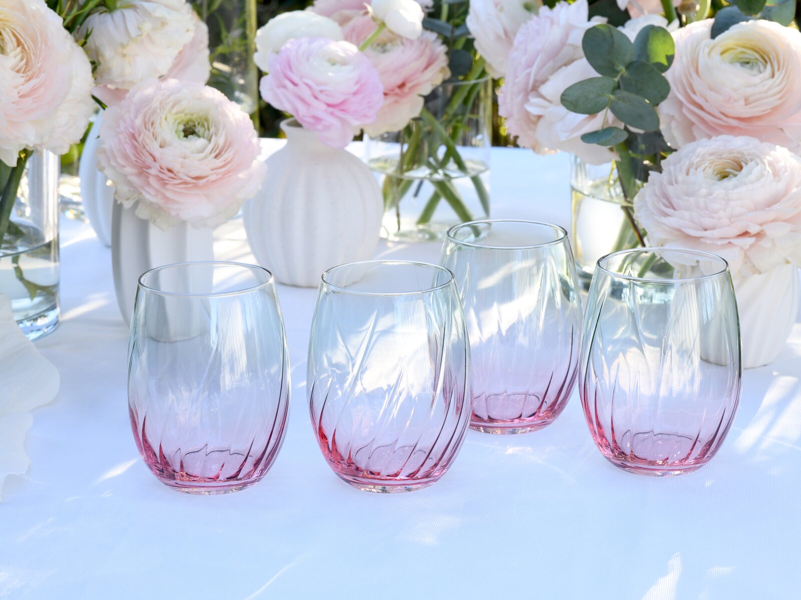 Vier mundgeblasene Trinkgläser ohne Stiel mit geriffelter Struktur und Farbverlauf von klar zu Rosa auf weißer Tischdecke, dekoriert mit zarten Blumen im Hintergrund; Perspektive: leicht von vorne und leicht von oben.