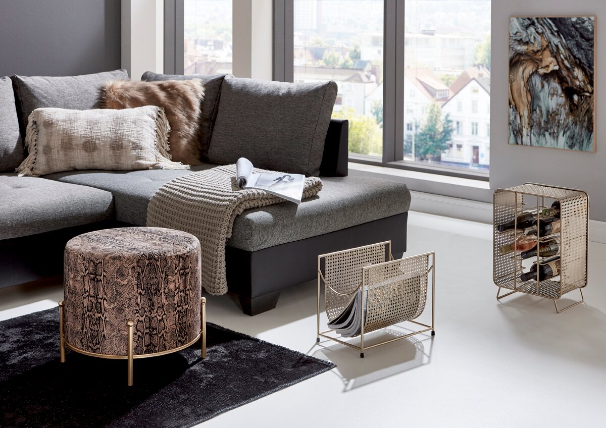 Modernes Wohnzimmer mit grauem Ecksofa, Schlangenmuster-Hocker, goldfarbenem Magazinhalter und Weinregal vor großen Fenstern; Perspektive: leicht erhöhte, schräge Frontansicht.
