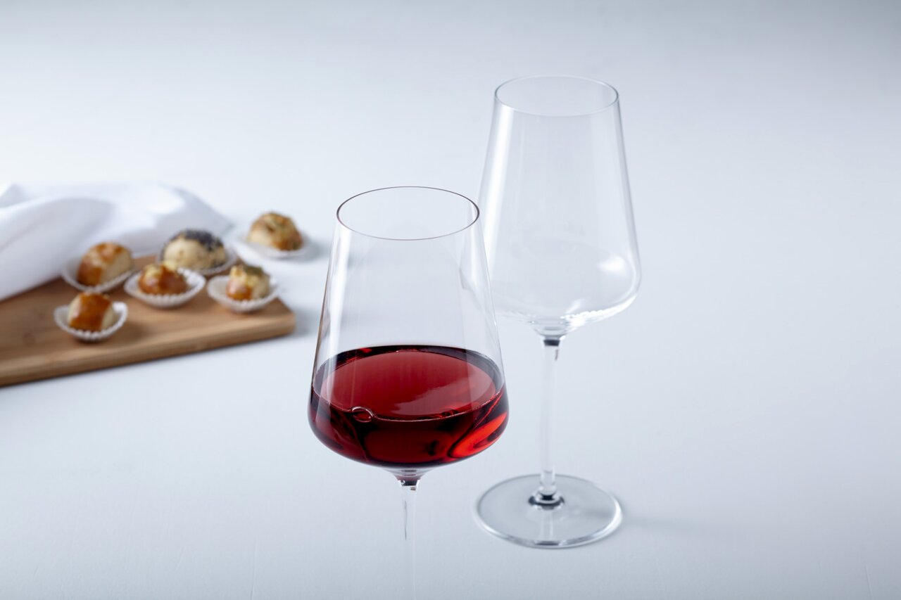 Zwei Rotweingläser Puccini 750ml, eines gefüllt mit Rotwein, auf einem Tisch mit kleinen Gebäckstücken im Hintergrund, seitliche Perspektive.