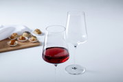 Twee Puccini-rodewijnglazen van 750 ml, waarvan één gevuld met rode wijn, op een tafel met kleine gebakjes op de achtergrond, gezien vanuit een zijdelingse hoek.
