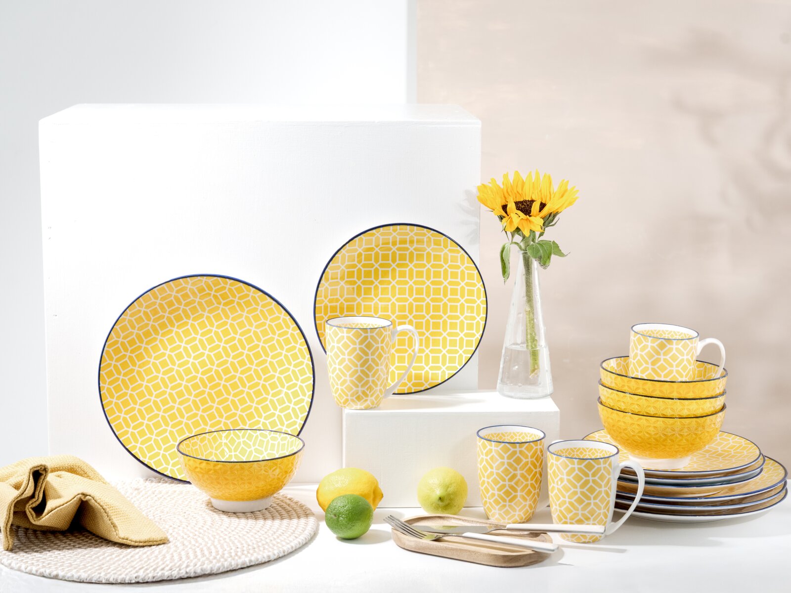 Creatable Kombiservice 16-tlg MEDITERRAN Gelbes Geschirrset mit geometrischem Muster und blauem Rand, bestehend aus Tellern, Schalen und Bechern, arrangiert auf weißen Podesten mit Sonnenblume und Zitrusfrüchten, Frontansicht in leicht schräger Perspektive.