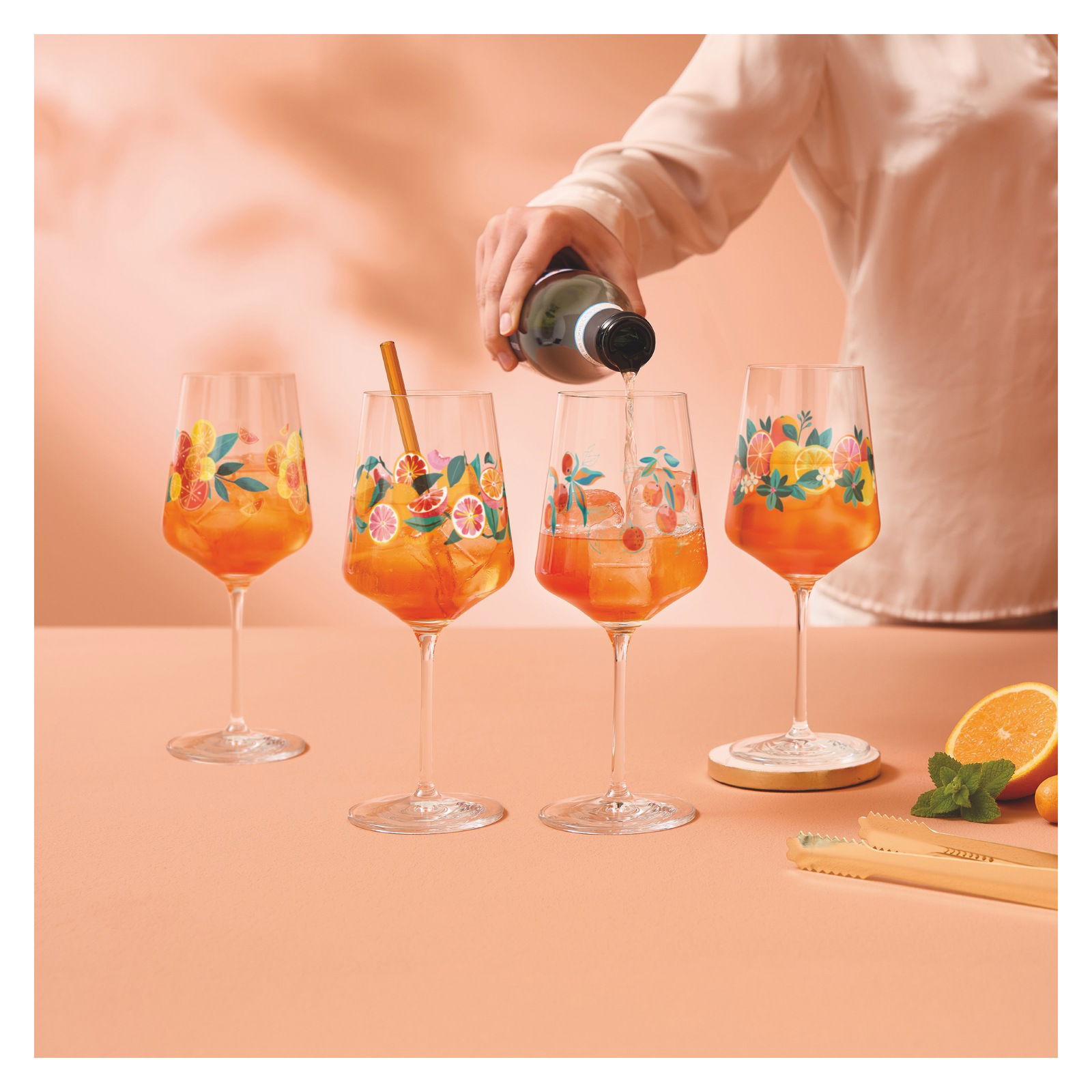 Ritzenhoff Aperizzo 4er-Set SOMMERRAUSCH Vier bunte Weingläser mit Fruchtmotiven, gefüllt mit einem orangen Getränk, auf einem Tisch. Eine Hand gießt ein Getränk in eines der Gläser. Perspektive von vorne.