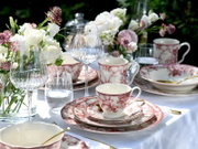 Festlich gedeckter Tisch mit weißem Tischtuch, rot-weiß gemustertem Porzellan (Tassen, Teller, Schalen), Weingläsern und Wassergläsern sowie Blumenarrangements; Perspektive: leicht erhöhte Frontansicht im Nahbereich.