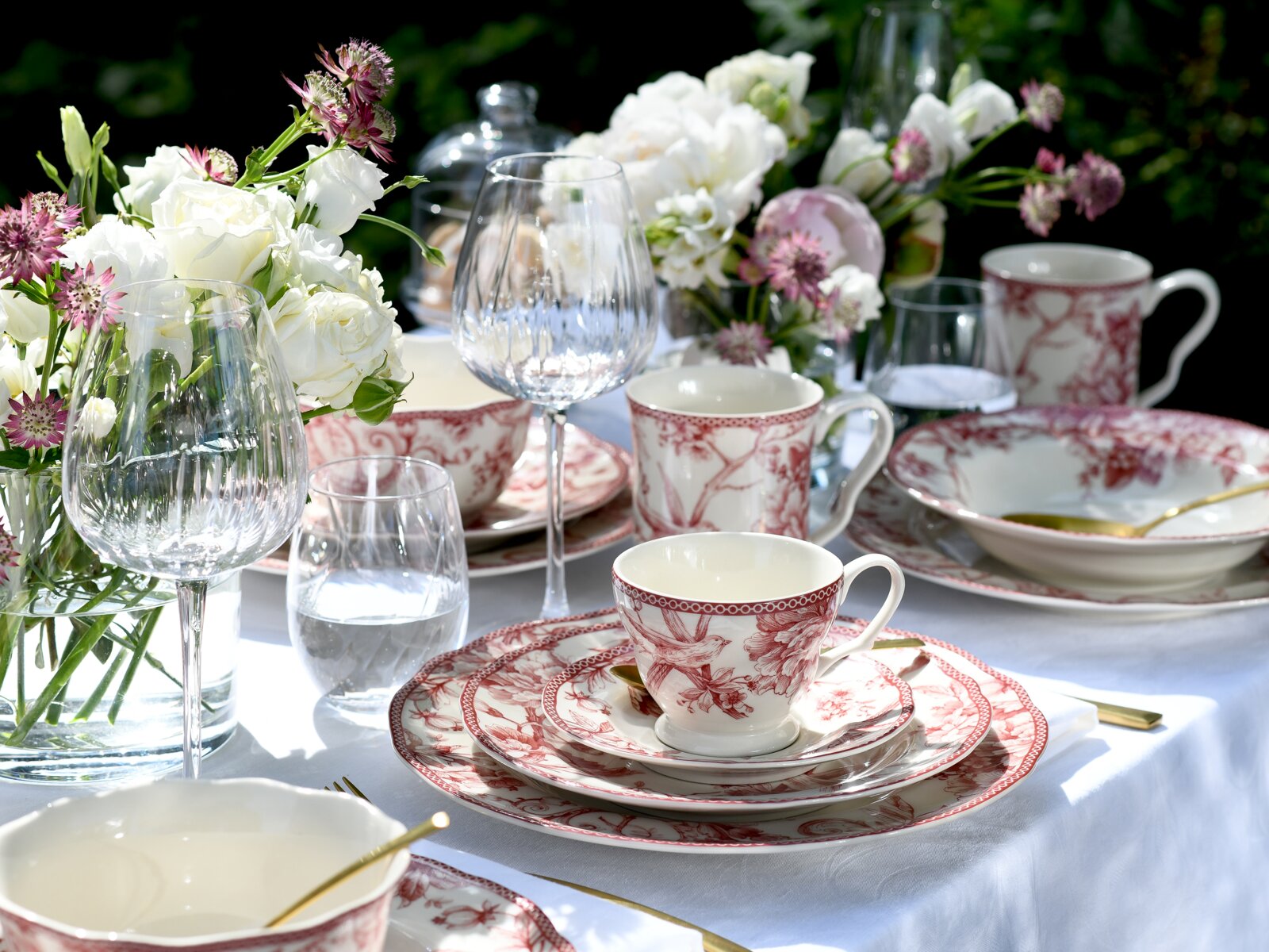 Festlich gedeckter Tisch mit weißem Tischtuch, rot-weiß gemustertem Porzellan (Tassen, Teller, Schalen), Weingläsern und Wassergläsern sowie Blumenarrangements; Perspektive: leicht erhöhte Frontansicht im Nahbereich.