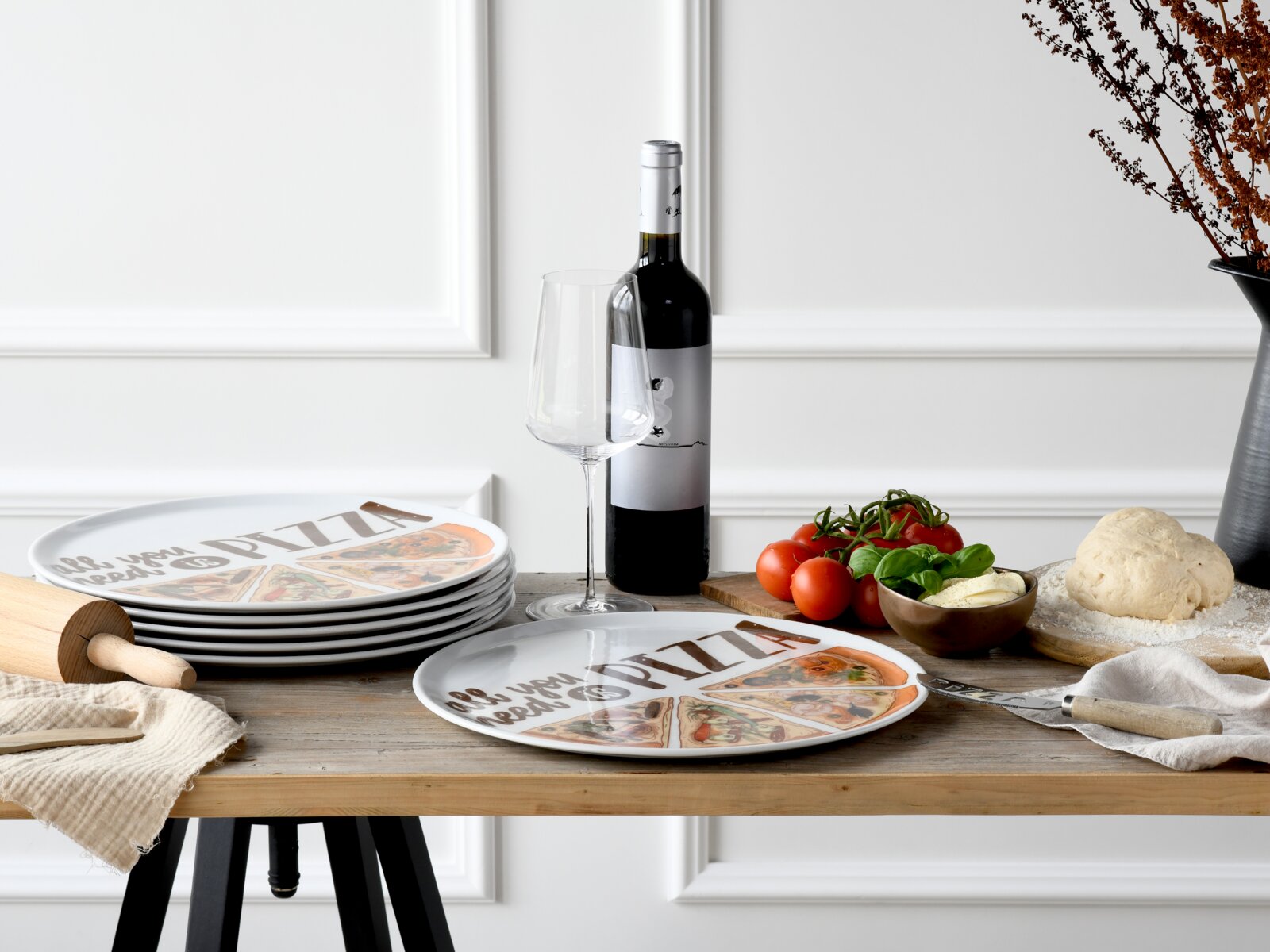 Runder Pizzateller mit Pizza-Illustrationen und Schriftzug „Pizza“, auf Holztisch arrangiert mit Tellerstapel, Weinflasche und Weinglas, Tomaten, Basilikum und Teig; Aufnahme in frontaler, leicht erhöhter Perspektive.