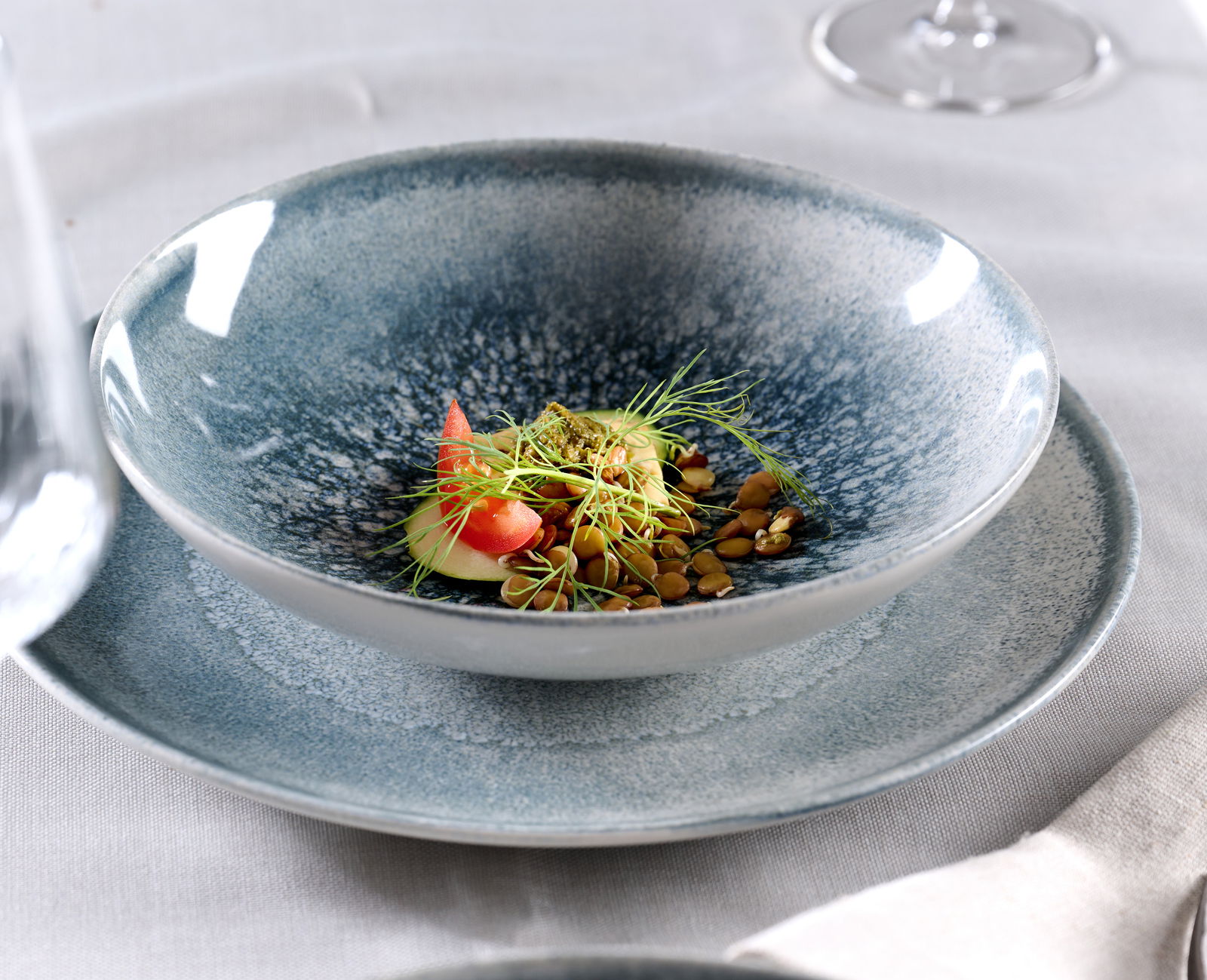 Nahaufnahme eines blauen Tellersets mit einem dekorativ angerichteten Gericht, bestehend aus Tomaten, Sprossen und Samen, auf einem Tisch mit weißer Tischdecke.