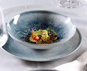 Close-up van een set blauwe borden met een decoratief opgemaakt gerecht, bestaande uit tomaten, spruitjes en zaden, op een tafel met een wit tafelkleed.