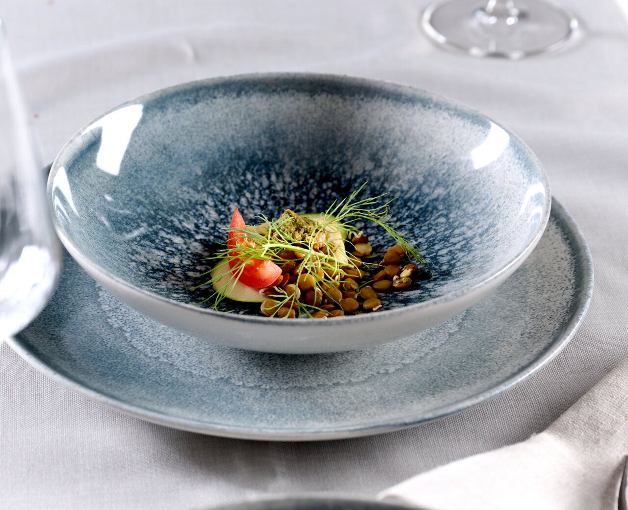 Close-up van een set blauwe borden met een decoratief opgemaakt gerecht, bestaande uit tomaten, spruitjes en zaden, op een tafel met een wit tafelkleed.